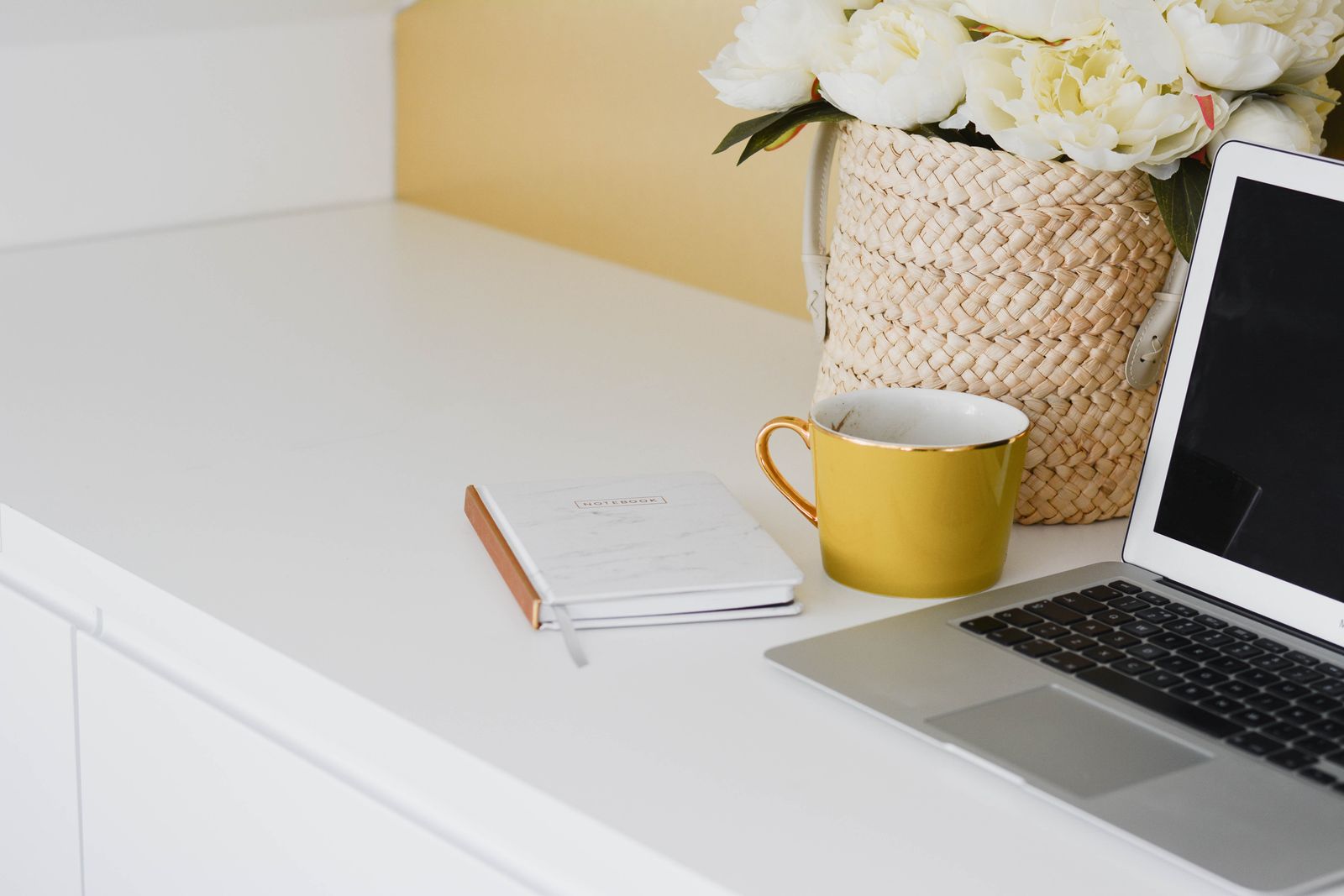 Ein weisser Schreibtisch mit geschlossenem Notizbuch, gelber Tasse, Blumenkorb und dem Rand eines Laptops.