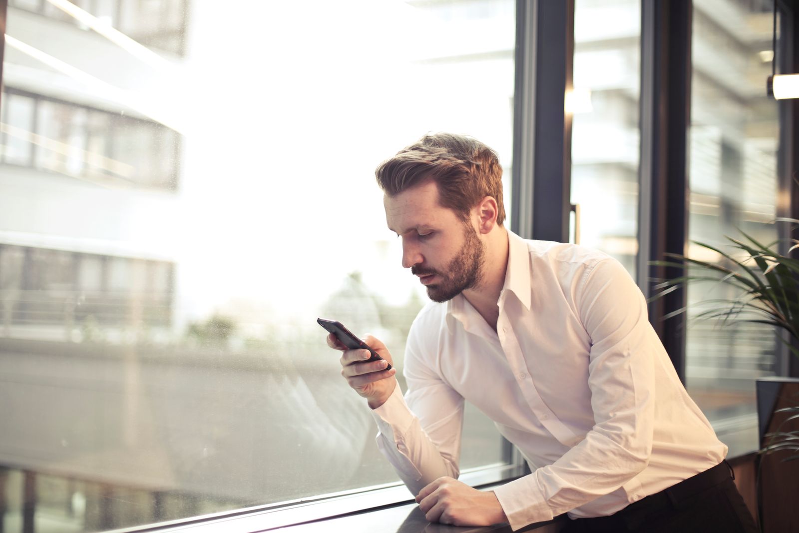 Ein Mann im weissen Hemd schaut neben einem grossen Bürofenster auf sein Telefon.