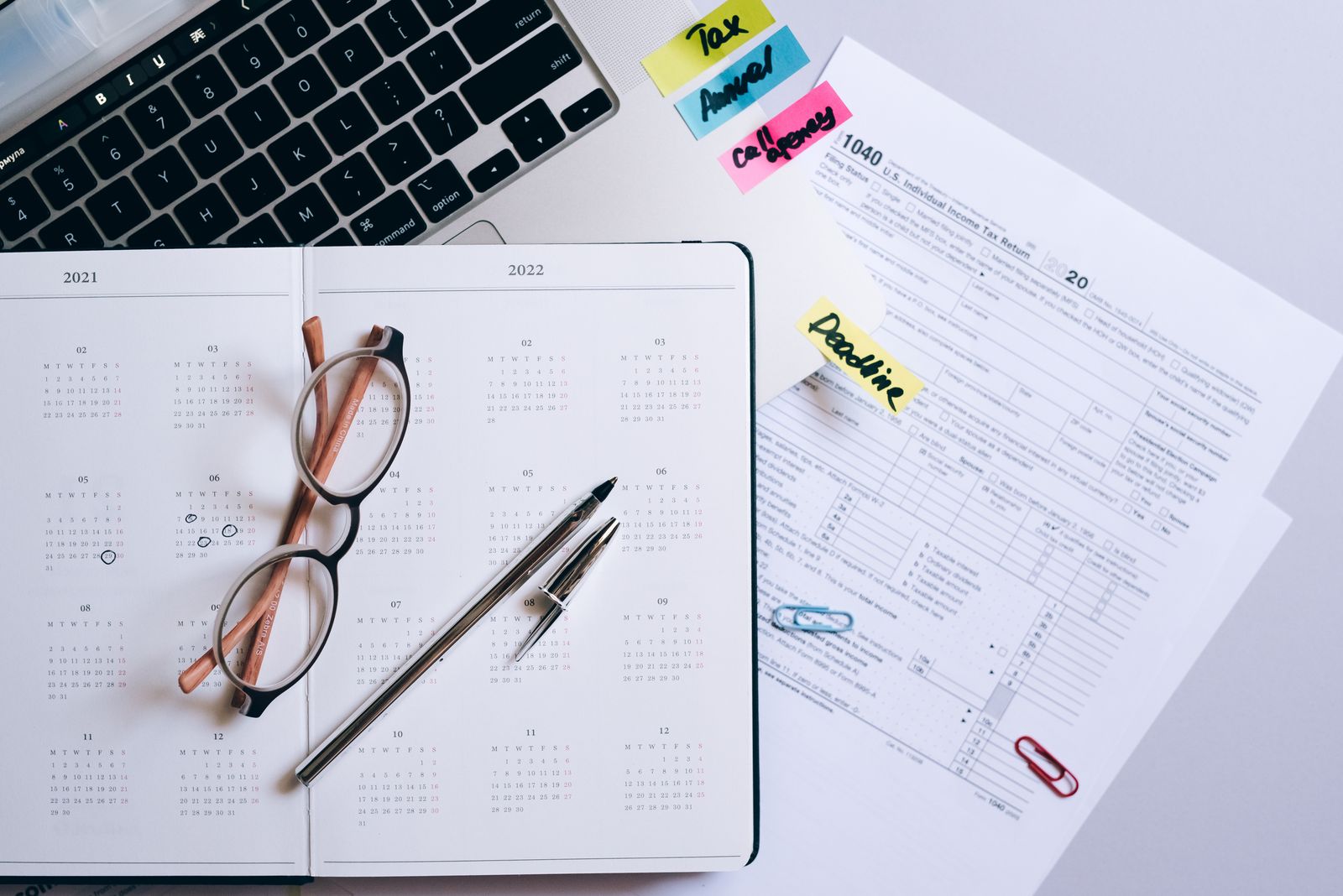 Flach fotografiert: ein Planer mit Brille und Stift, ein Steuerformular und Haftnotizen neben einer Laptop-Tastatur.