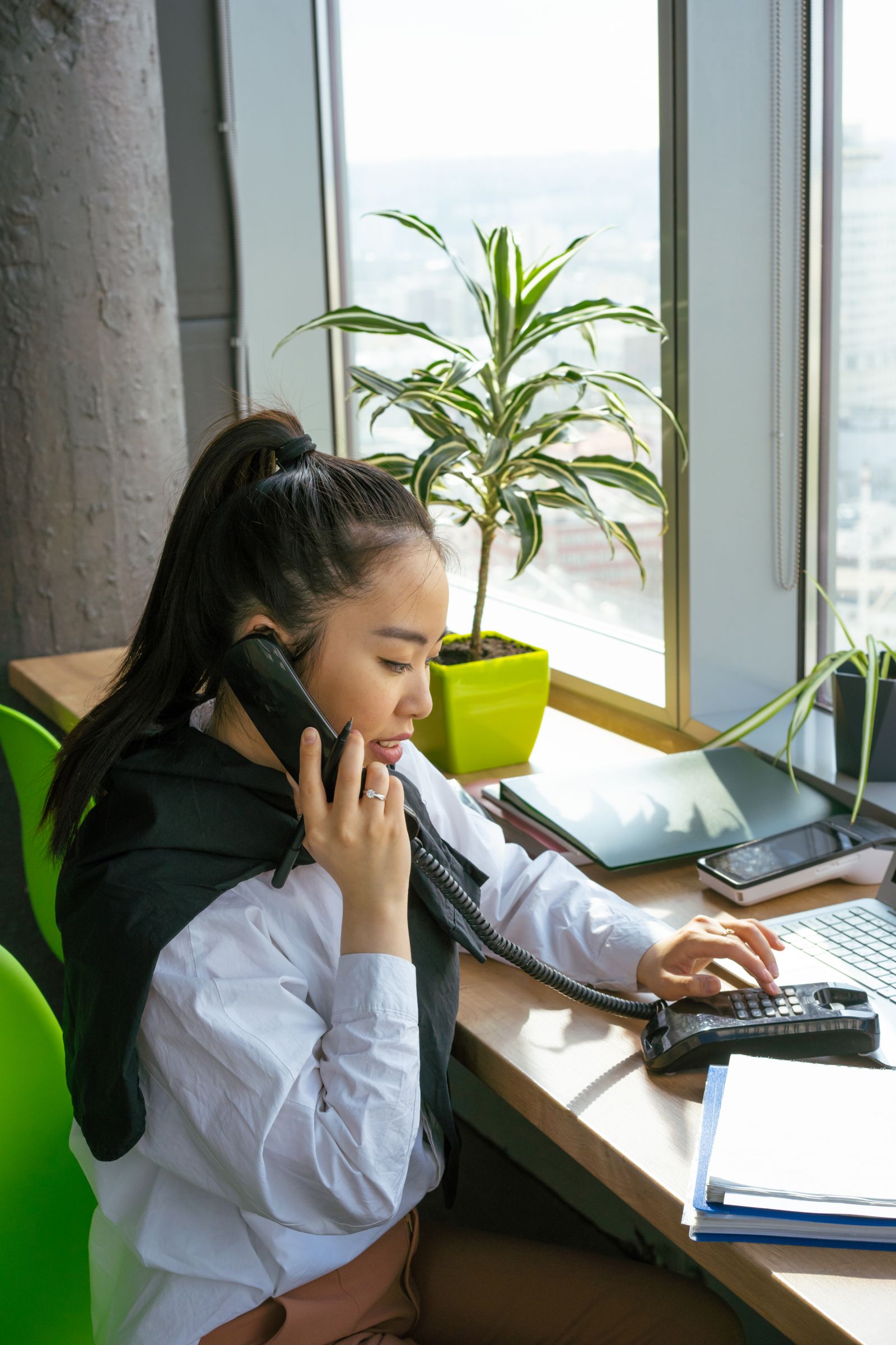 Eine Frau am Fensterplatz telefoniert mit einem Festnetztelefon und drückt die Tasten, umgeben von Pflanzen und Ordnern.